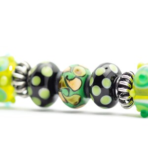 May include: A close-up of a bracelet with black, green, and yellow glass beads. The beads have different patterns, including polka dots, swirls, and abstract designs.