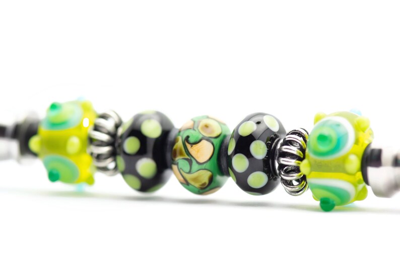 May include: A close-up of a bracelet with black, green, and yellow glass beads. The beads have different patterns, including polka dots, swirls, and abstract designs.