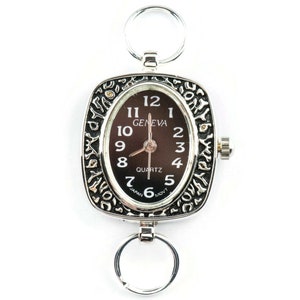 K&ouml;nnte beinhalten: Eine silberne Taschenuhr mit schwarzem Zifferblatt und verziertem silbernen Rand. Das Zifferblatt der Uhr hat schwarze r&ouml;mische Ziffern als Stundenmarkierungen und die Worte "GENEVA QUARTZ" sind darauf gedruckt.