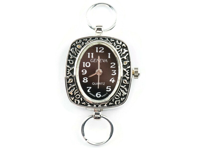 K&ouml;nnte beinhalten: Eine silberne Taschenuhr mit schwarzem Zifferblatt und verziertem silbernen Rand. Das Zifferblatt der Uhr hat schwarze r&ouml;mische Ziffern als Stundenmarkierungen und die Worte "GENEVA QUARTZ" sind darauf gedruckt.