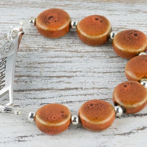May include: A silver medical alert bracelet with the word "ASTHMA" engraved on it. The bracelet has a clasp and is adorned with orange ceramic beads.