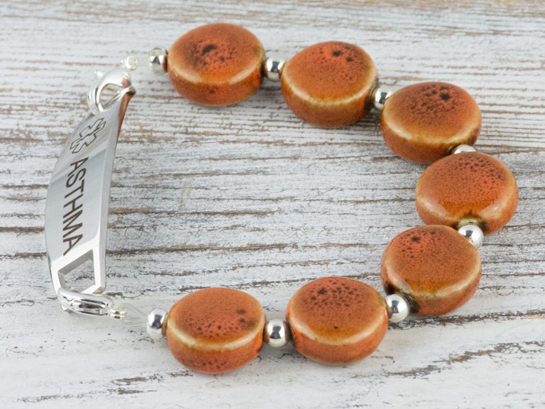 May include: A silver medical alert bracelet with the word "ASTHMA" engraved on it. The bracelet has a clasp and is adorned with orange ceramic beads.