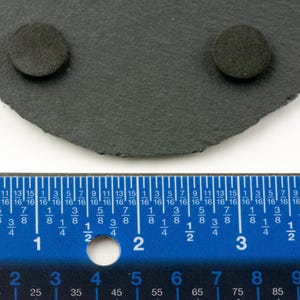May include: A dark gray, irregularly shaped slate coaster with two small, round, dark gray pads attached. A blue ruler with inch and centimeter markings is in the foreground, showing measurements.