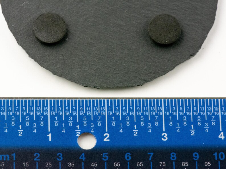 May include: A dark gray, irregularly shaped slate coaster with two small, round, dark gray pads attached. A blue ruler with inch and centimeter markings is in the foreground, showing measurements.