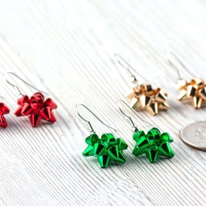 May include: A collection of bow-shaped earrings in red, green, and gold. The earrings are on silver hooks and are arranged on a white wooden surface. A US quarter is next to the earrings.