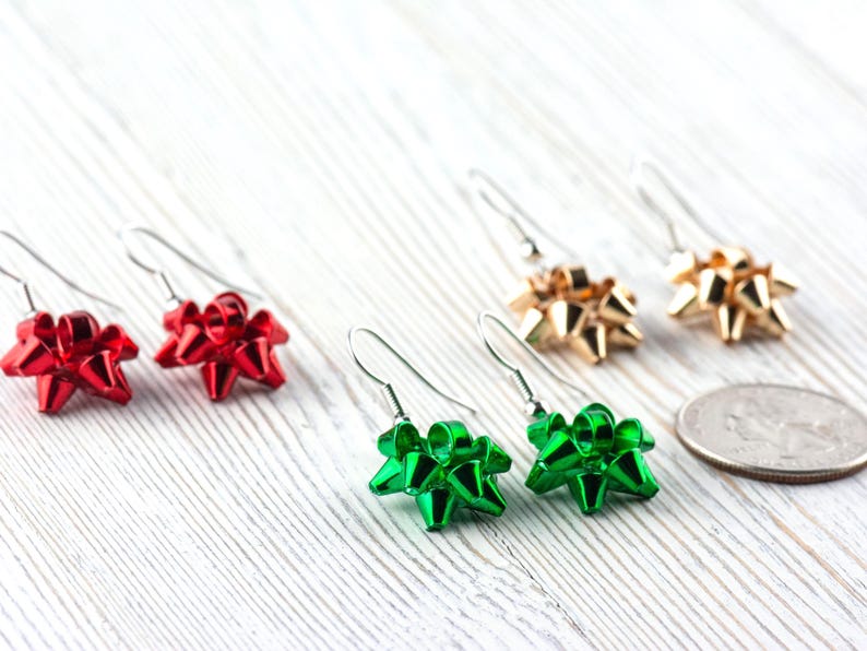 May include: A collection of bow-shaped earrings in red, green, and gold. The earrings are on silver hooks and are arranged on a white wooden surface. A US quarter is next to the earrings.
