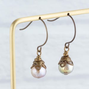 May include: A pair of antique-style earrings with pear-shaped pearls. The earrings have bronze-colored ear wires and decorative caps. The pearls have a soft, iridescent sheen, and the earrings are displayed on a gold-colored stand.