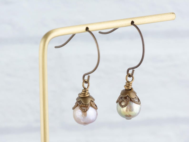 May include: A pair of antique-style earrings with pear-shaped pearls. The earrings have bronze-colored ear wires and decorative caps. The pearls have a soft, iridescent sheen, and the earrings are displayed on a gold-colored stand.