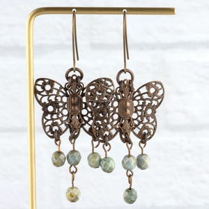May include: Ornate bronze-colored butterfly earrings with intricate cut-out designs. Each earring features three dangling, faceted, pale green beads. The earrings are displayed on a gold-colored stand against a white brick background.