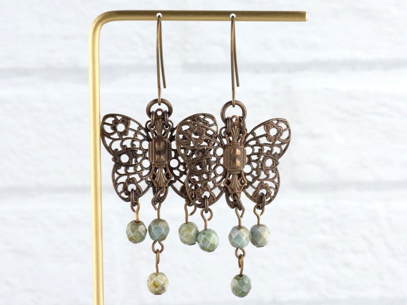 May include: Ornate bronze-colored butterfly earrings with intricate cut-out designs. Each earring features three dangling, faceted, pale green beads. The earrings are displayed on a gold-colored stand against a white brick background.