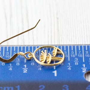 May include: A gold-colored earring with a circular design featuring a stylized tree silhouette. The earring is measured against a blue ruler with inch markings.
