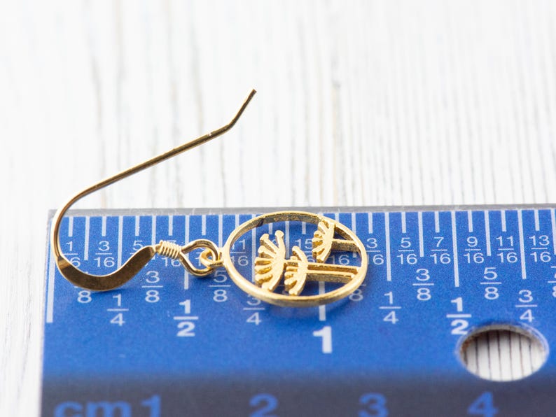 May include: A gold-colored earring with a circular design featuring a stylized tree silhouette. The earring is measured against a blue ruler with inch markings.
