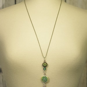 May include: A silver chain necklace with a green and brown pendant featuring a floral design. The pendant is attached to a pink tassel.