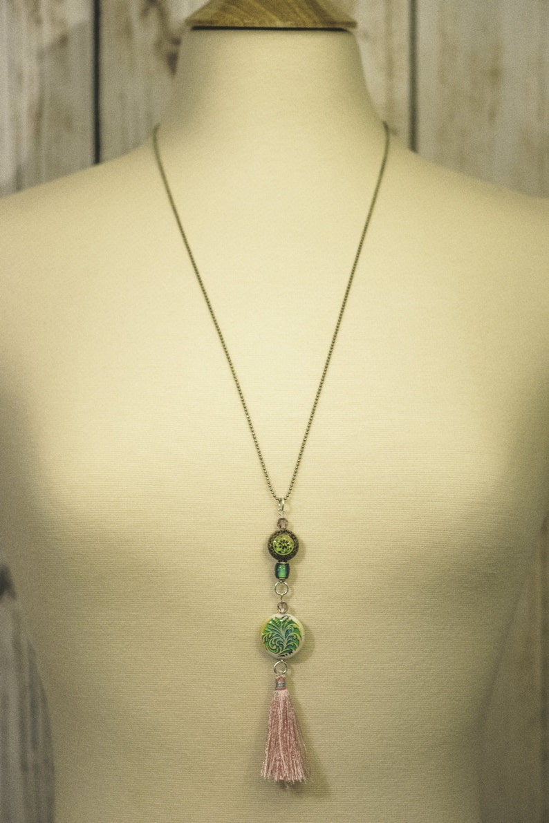 May include: A silver chain necklace with a green and brown pendant featuring a floral design. The pendant is attached to a pink tassel.