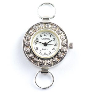Pu&ograve; includere: Un orologio al quarzo Geneva argentato con quadrante bianco e lancette nere. L'orologio ha un design circolare con un anello nella parte superiore e inferiore per il fissaggio. Il quadrante &egrave; circondato da un bordo decorativo con perline.