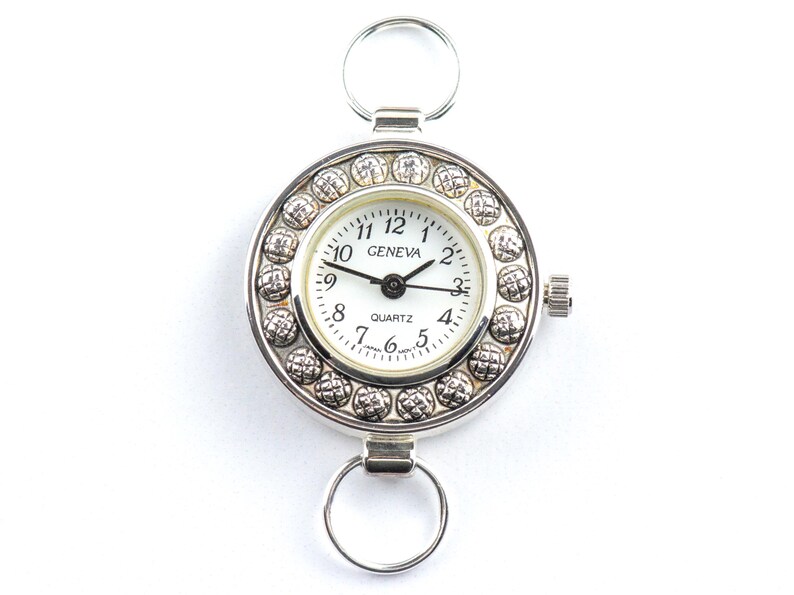 Pu&ograve; includere: Un orologio al quarzo Geneva argentato con quadrante bianco e lancette nere. L'orologio ha un design circolare con un anello nella parte superiore e inferiore per il fissaggio. Il quadrante &egrave; circondato da un bordo decorativo con perline.