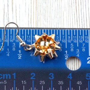 May include: A single gold-colored bow earring with a silver hook. The bow is approximately 1 inch in diameter. The earring is set against a blue ruler with both inch and centimeter measurements.