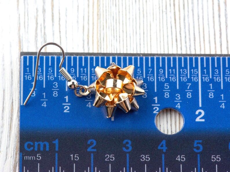 May include: A single gold-colored bow earring with a silver hook. The bow is approximately 1 inch in diameter. The earring is set against a blue ruler with both inch and centimeter measurements.