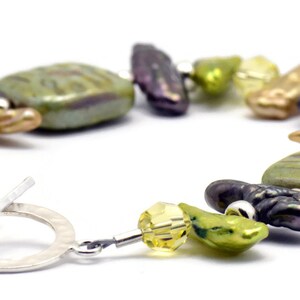 K&ouml;nnte beinhalten: Ein Armband aus S&uuml;&szlig;wasserperlen in Gr&uuml;n, Braun und Gold mit einem silbernen Schiebeverschluss. Das Armband verf&uuml;gt &uuml;ber eine Vielzahl von Formen und Gr&ouml;&szlig;en von Perlen, darunter runde, ovale und quadratische.