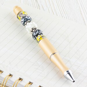 May include: A gold pen with black, white, and yellow glass beads. The pen is lying on a lined notebook.