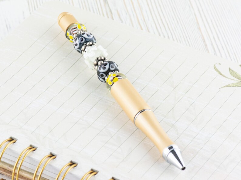 May include: A gold pen with black, white, and yellow glass beads. The pen is lying on a lined notebook.
