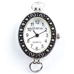 May include: Oval Geneva quartz watch with a white face, black hands, and black numerals. The watch has a silver-colored, ornate bezel with a decorative pattern. The watch has a loop at the top and bottom for attachment.