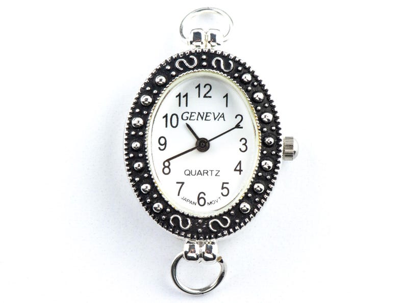 May include: Oval Geneva quartz watch with a white face, black hands, and black numerals. The watch has a silver-colored, ornate bezel with a decorative pattern. The watch has a loop at the top and bottom for attachment.