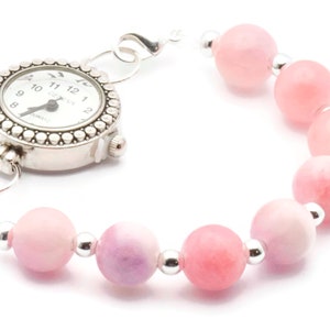 Puede incluir: Pulsera de reloj plateada con esfera blanca y cuentas de piedras preciosas rosas. La esfera tiene n&uacute;meros y manecillas negras. La pulsera presenta cuentas redondas de color rosa claro separadas por peque&ntilde;as cuentas plateadas.