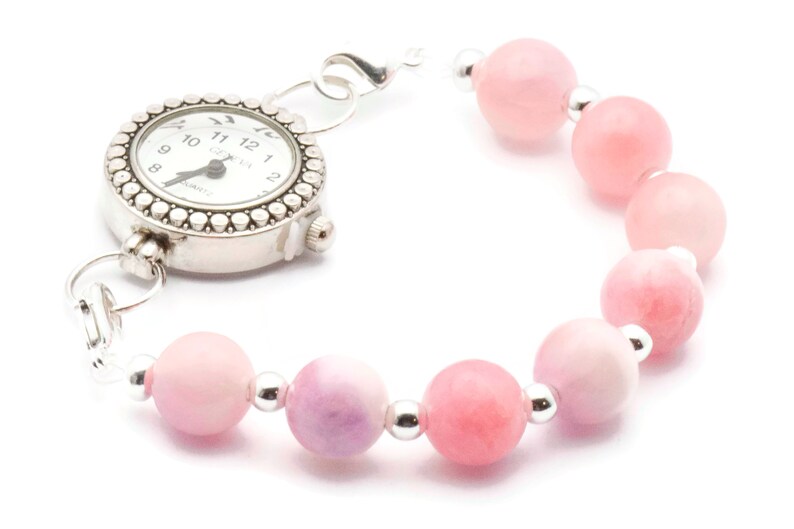 Puede incluir: Pulsera de reloj plateada con esfera blanca y cuentas de piedras preciosas rosas. La esfera tiene n&uacute;meros y manecillas negras. La pulsera presenta cuentas redondas de color rosa claro separadas por peque&ntilde;as cuentas plateadas.