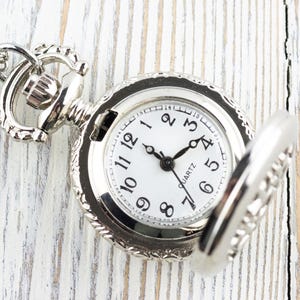 May include: A silver pocket watch with a white face and black hands. The watch has a chain attached to it and is open to reveal the inner workings.
