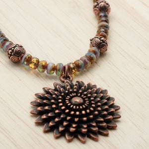 Puede incluir: Un collar de cobre con un gran colgante de cobre en forma de flor adornada. El collar est&aacute; hecho con cuentas de vidrio iridiscente y separadores de cobre.