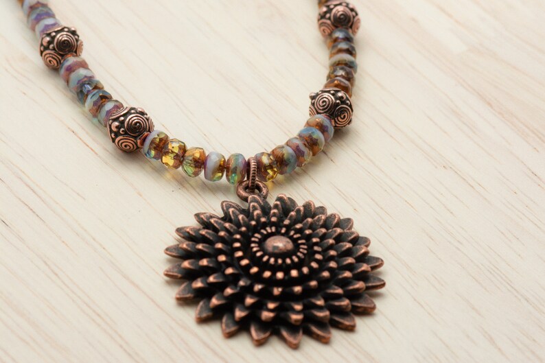 Puede incluir: Un collar de cobre con un gran colgante de cobre en forma de flor adornada. El collar est&aacute; hecho con cuentas de vidrio iridiscente y separadores de cobre.