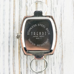 Puede incluir: Un reloj rectangular con parte trasera de acero inoxidable, con el texto "TRENDZ JAPAN MOVT BSR-1 WATER RESISTANT" impreso en la esfera. El reloj tiene un bisel plateado y un anillo plateado en la parte superior e inferior.