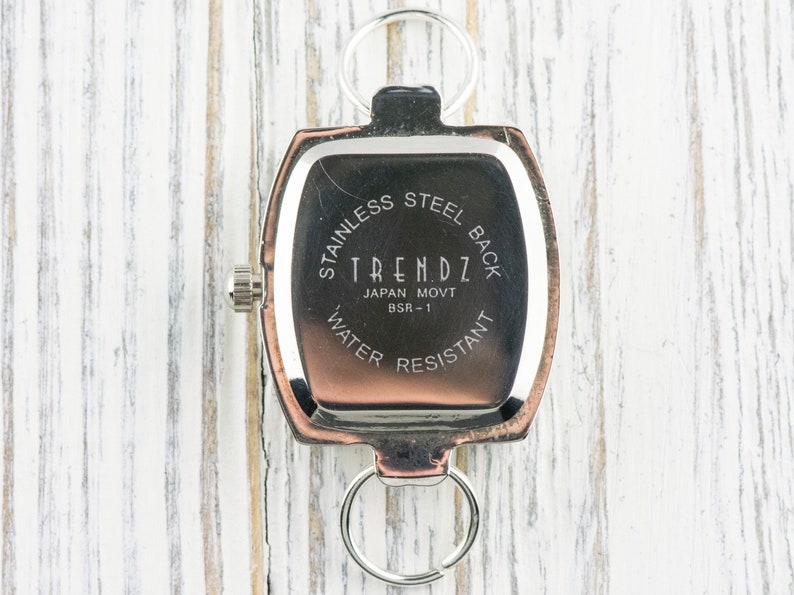 Puede incluir: Un reloj rectangular con parte trasera de acero inoxidable, con el texto "TRENDZ JAPAN MOVT BSR-1 WATER RESISTANT" impreso en la esfera. El reloj tiene un bisel plateado y un anillo plateado en la parte superior e inferior.