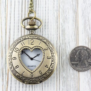 Pode incluir: Um rel&oacute;gio de bolso dourado com uma janela em forma de cora&ccedil;&atilde;o que revela o movimento de quartzo. O rel&oacute;gio tem numerais romanos e uma corrente anexa.