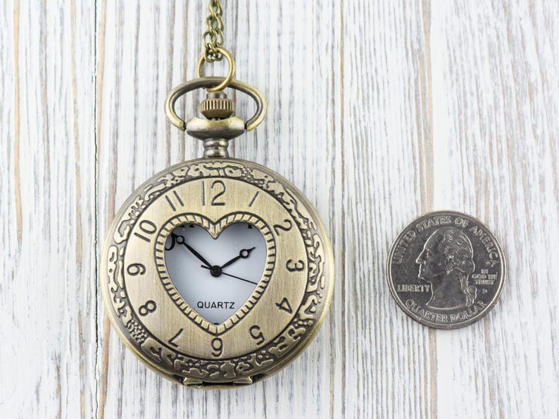 Pode incluir: Um rel&oacute;gio de bolso dourado com uma janela em forma de cora&ccedil;&atilde;o que revela o movimento de quartzo. O rel&oacute;gio tem numerais romanos e uma corrente anexa.