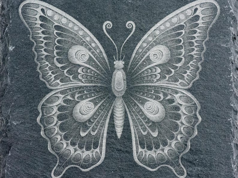 Puede incluir: Un dise&ntilde;o de mariposa detallado e intrincado tallado en una superficie de pizarra gris oscuro. La mariposa es blanca y tiene un patr&oacute;n sim&eacute;trico de c&iacute;rculos y espirales.