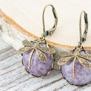Pode incluir: Um par de brincos pendentes com uma pedra de &aacute;gata roxa e um amuleto de lib&eacute;lula dourado.