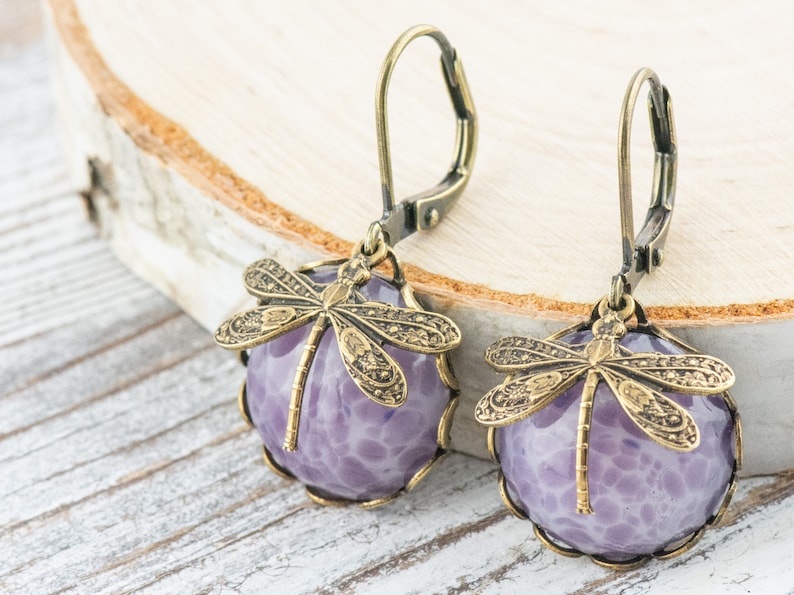 Pode incluir: Um par de brincos pendentes com uma pedra de &aacute;gata roxa e um amuleto de lib&eacute;lula dourado.