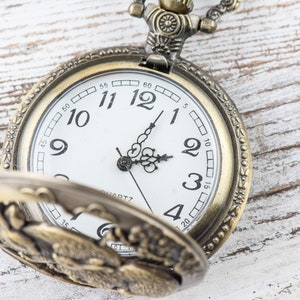 Puede incluir: Reloj de bolsillo antiguo de lat&oacute;n con esfera blanca, n&uacute;meros romanos y manecillas negras. La tapa del reloj est&aacute; abierta, revelando su intrincado dise&ntilde;o. La hora es aproximadamente las 2:05. La palabra "QUARTZ" est&aacute; impresa en la esfera.