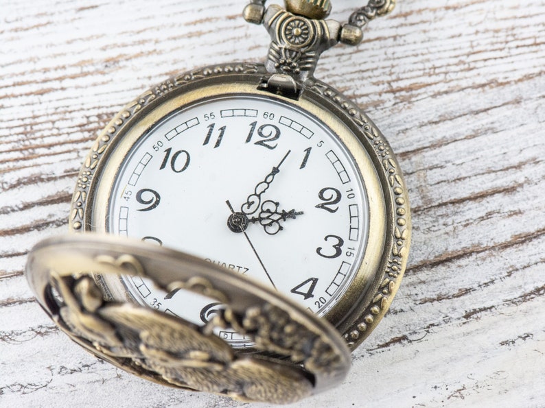 Puede incluir: Reloj de bolsillo antiguo de lat&oacute;n con esfera blanca, n&uacute;meros romanos y manecillas negras. La tapa del reloj est&aacute; abierta, revelando su intrincado dise&ntilde;o. La hora es aproximadamente las 2:05. La palabra "QUARTZ" est&aacute; impresa en la esfera.