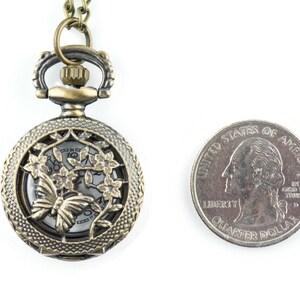 May include: A bronze pocket watch necklace with a delicate butterfly and floral design. The watch face is visible through the intricate cut-out design. The necklace is shown next to a US quarter for size comparison.