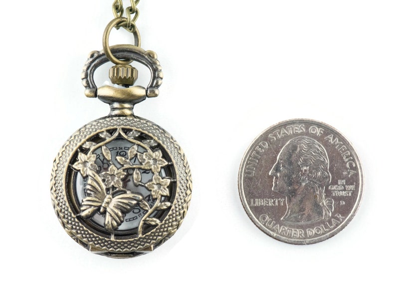 May include: A bronze pocket watch necklace with a delicate butterfly and floral design. The watch face is visible through the intricate cut-out design. The necklace is shown next to a US quarter for size comparison.