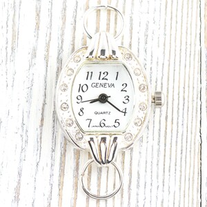May include: Silver-toned Geneva quartz watch with a white face and black hands. The watch has a decorative bezel with clear rhinestones and a small crown. The watch face displays the numbers 1 through 12. The watch has two loops for attaching to a chain.