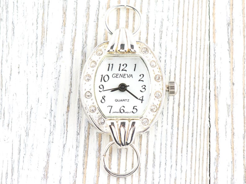 May include: Silver-toned Geneva quartz watch with a white face and black hands. The watch has a decorative bezel with clear rhinestones and a small crown. The watch face displays the numbers 1 through 12. The watch has two loops for attaching to a chain.