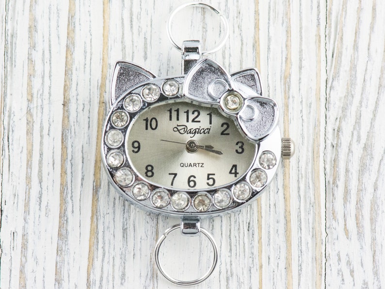 May include: A silver-toned watch with a white face and rhinestone accents. The watch is shaped like a cat's head with a bow on top. The watch face reads "Dagicci Quartz".