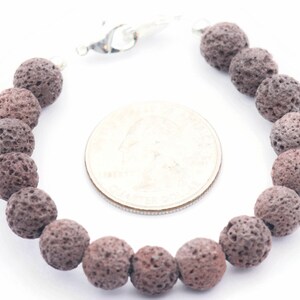 Puede incluir: Un brazalete con cuentas de roca volc&aacute;nica gris oscuro y porosa. Las cuentas redondas est&aacute;n ensartadas con un cierre plateado. Una moneda est&aacute; al lado del brazalete para la comparaci&oacute;n de tama&ntilde;os.