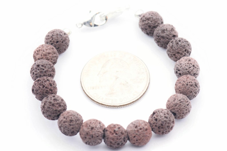 Puede incluir: Un brazalete con cuentas de roca volc&aacute;nica gris oscuro y porosa. Las cuentas redondas est&aacute;n ensartadas con un cierre plateado. Una moneda est&aacute; al lado del brazalete para la comparaci&oacute;n de tama&ntilde;os.