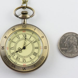 Op de afbeelding: Antieke bronzen zakhorloge met Romeinse cijfers op een cr&egrave;mekleurige wijzerplaat. Een Amerikaanse kwart dollar munt ligt ernaast ter vergelijking. Het horloge heeft een ketting en een decoratieve bovenkant.