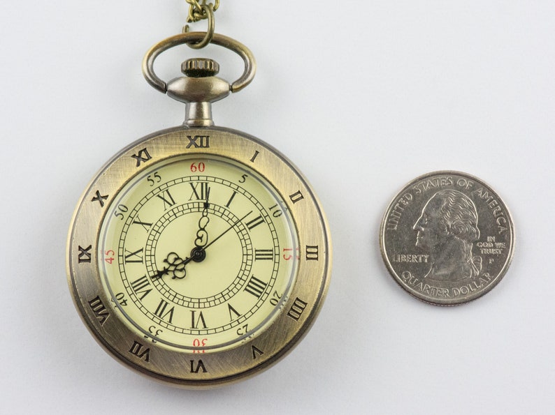 Op de afbeelding: Antieke bronzen zakhorloge met Romeinse cijfers op een cr&egrave;mekleurige wijzerplaat. Een Amerikaanse kwart dollar munt ligt ernaast ter vergelijking. Het horloge heeft een ketting en een decoratieve bovenkant.